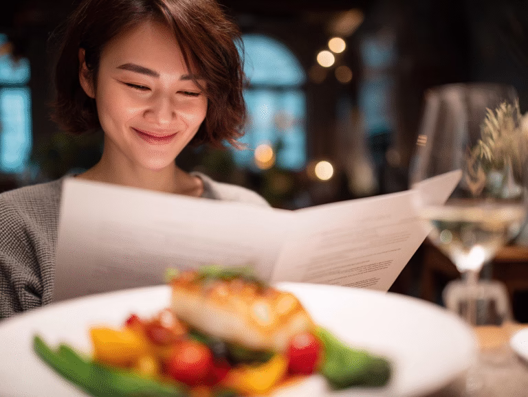 A smiling woman reads a restaurant menu with a plate of grilled fish and vegetables in front of her