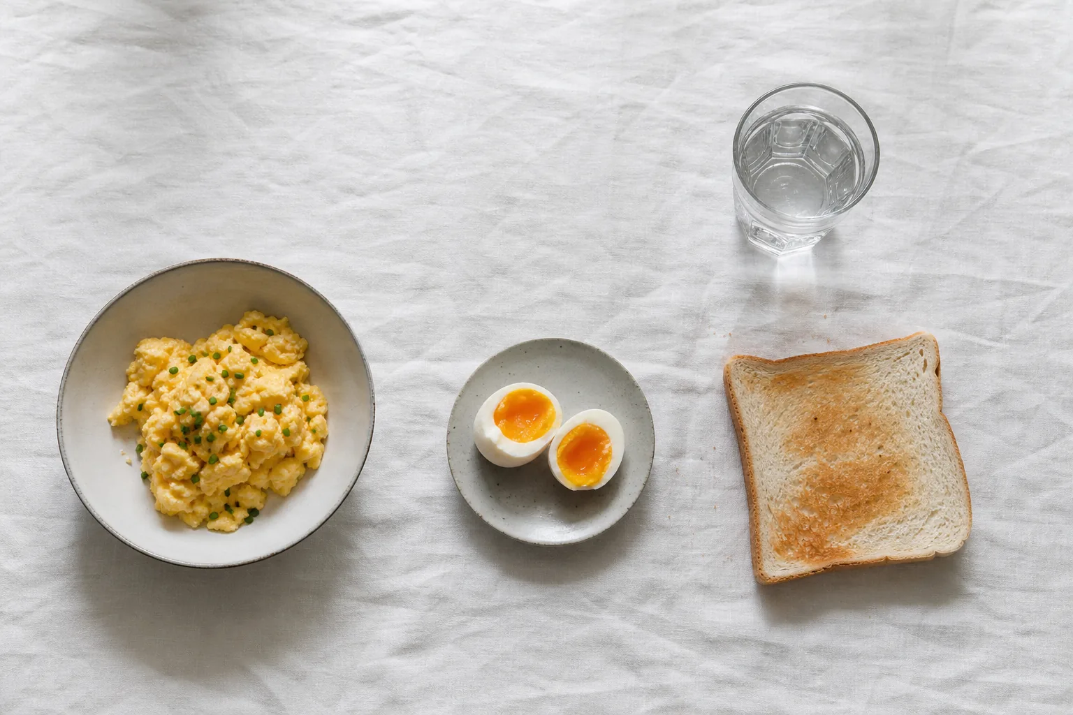 Soft scrambled eggs, a halved soft-boiled egg, plain white toast and a glass of water on white linen