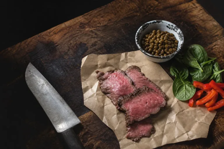 Slices of rare beef on butcher paper, a chipped enamel bowl of lentils, spinach leaves and red pepper strips on a block.