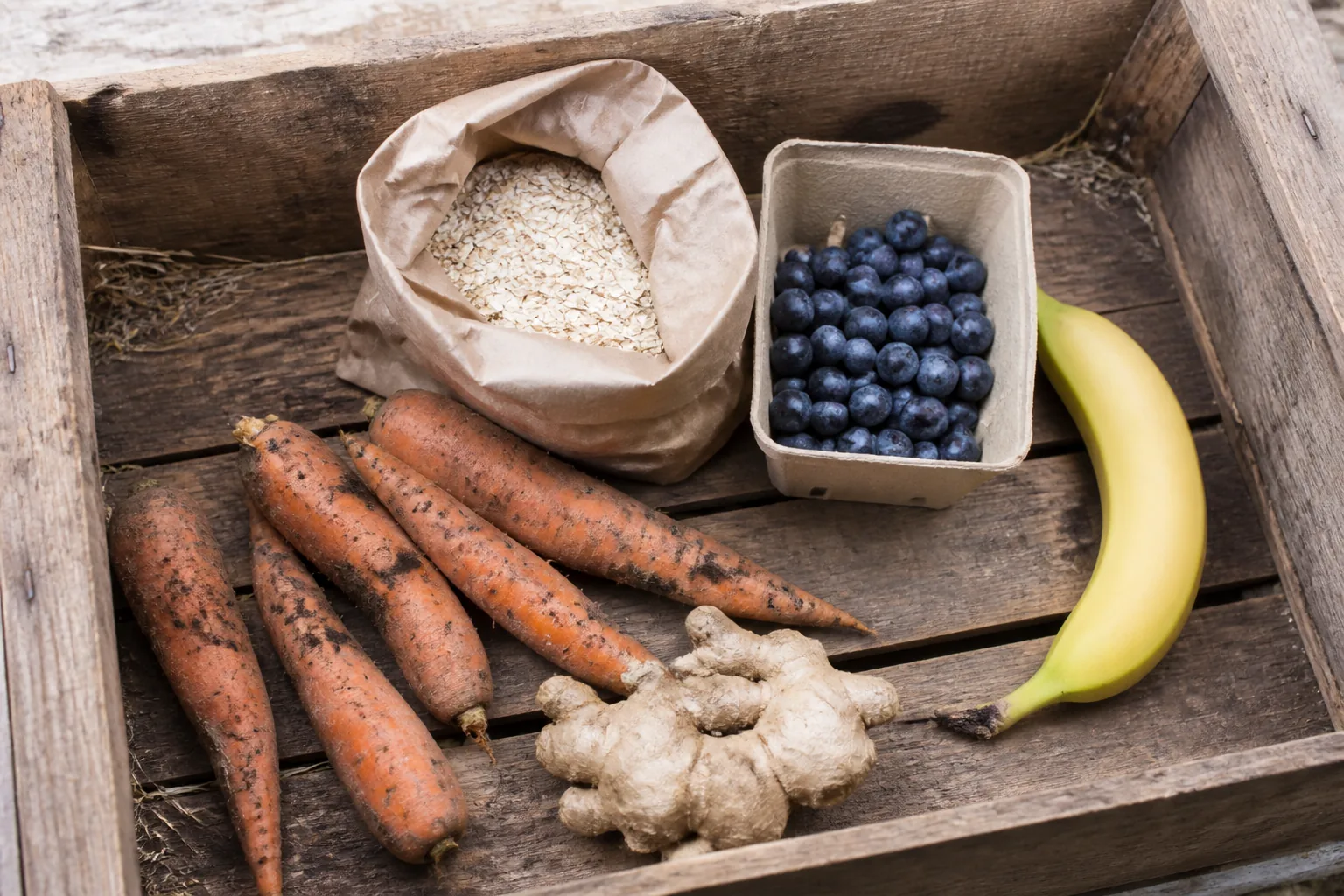 Produce in a wooden market crate: unwashed carrots, a paper bag of oats, blueberries, fresh ginger and a firm banana.